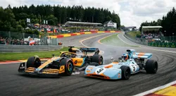 A modern papaya-orange McLaren racing wheel to wheel with a classic 1970s Gulf-liveried Formula 1 car through Spa-Francorchamps